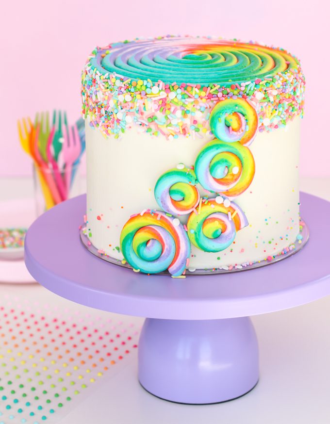 Rainbow swirl cake on purple cake stand with sprinkles.